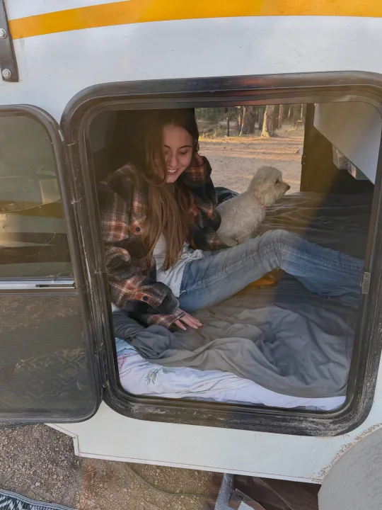 A smiling woman and her fluffy white dog relaxing inside a cozy RV rental. Perfect for outdoor adventures.. Lil’ Guy RET 2006