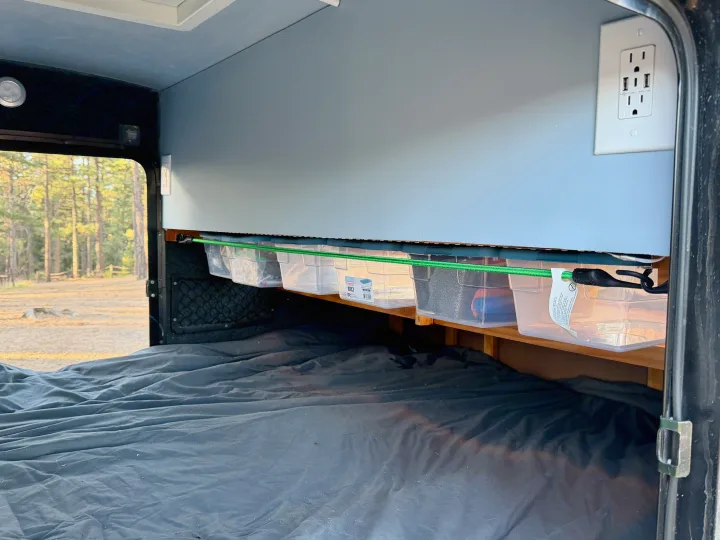 RV rental bedroom with dark bed, overhead storage bins, and power outlet. A green bungee secures bins. Forest outside window.. Lil’ Guy RET 2006