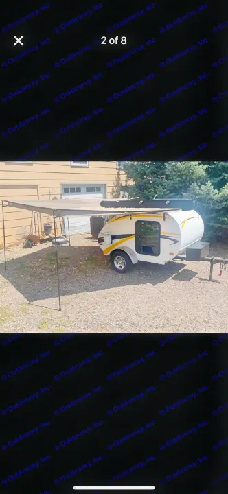 Compact teardrop camper with an awning, ideal for an rv rental adventure. Parked on gravel next to a house.. Lil’ Guy RET 2006