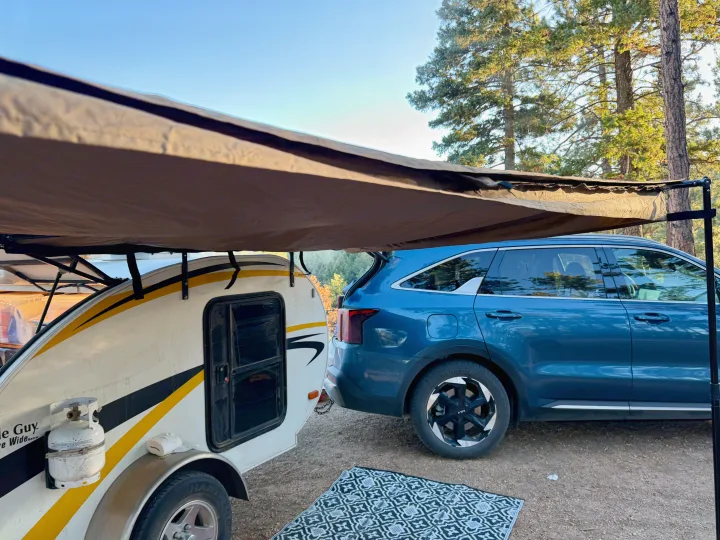 An RV rental teardrop camper with an awning extended, parked beside a blue SUV in a wooded campsite, ready for adventure.. Lil’ Guy RET 2006