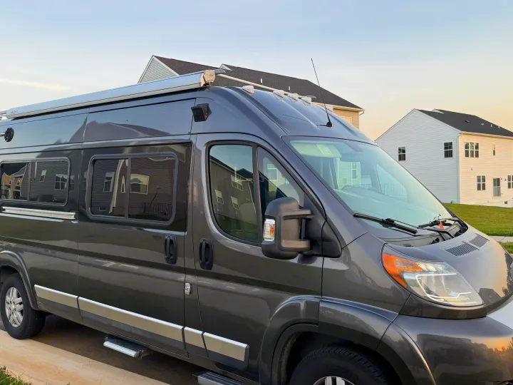 Gray camper van rv rental, ready for travel. Shows roof awning, side steps, and tinted windows in a residential setting.. Winnebago Travato 2016