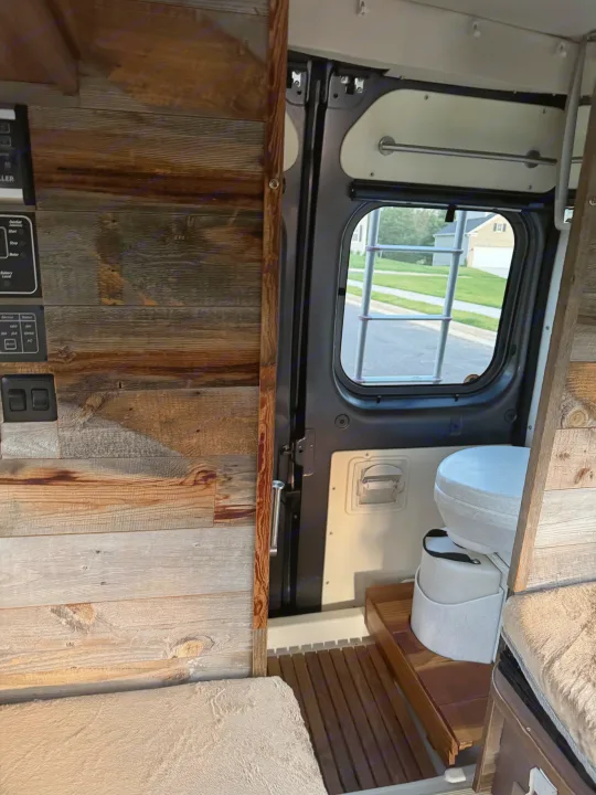 Explore this RV rental! Rustic wood interior, control panel, portable toilet, and cozy cushioned seating in a functional camper van setup.. Winnebago Travato 2016