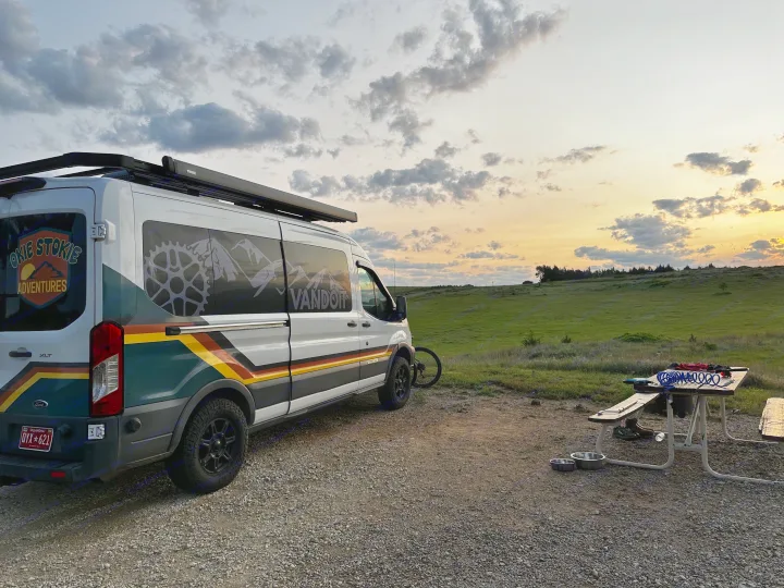 Camper van rental under a vibrant sunset with a custom-designed van, parked on gravel next to a picnic table in a grassy field.. Ford Transit 2018