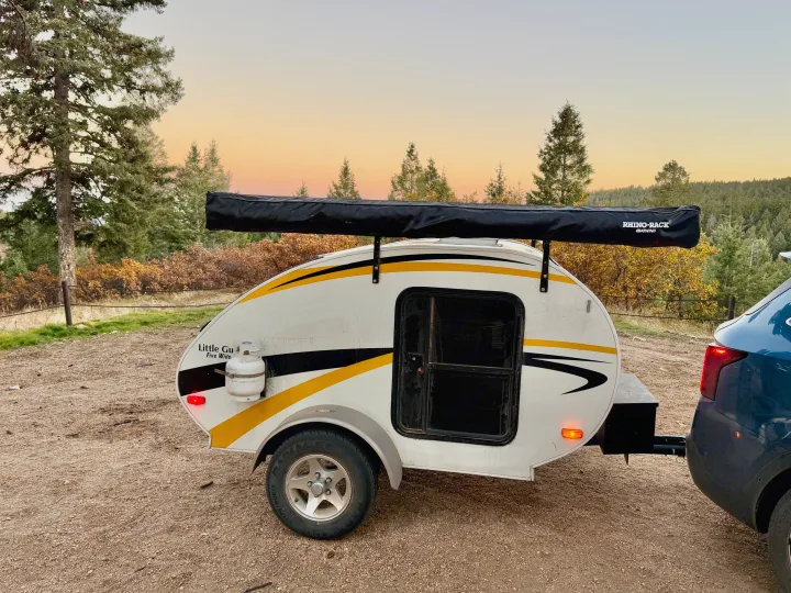 White teardrop camper RV rental with a side awning, hitched to a car in a scenic, tree-filled landscape at sunset.. Lil’ Guy RET 2006