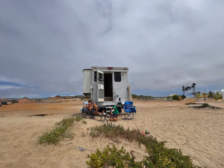 A family enjoys their RV rental on a sandy beach. A man and child relax in chairs outside their camper on an overcast day.. Ford F350 Diesel Double Cab 2017