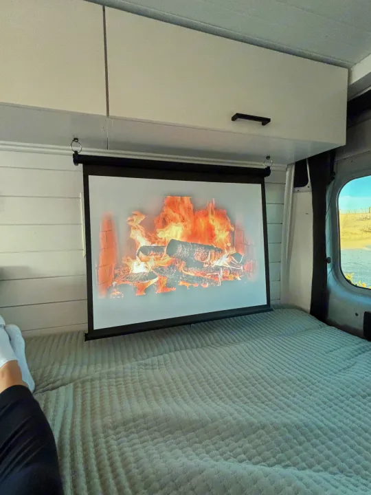 Cozy rv rental interior: fireplace projected on a screen above a bed. A person relaxes with feet up, enjoying the van life ambiance.. Ram Ram ProMaster 2500 2019