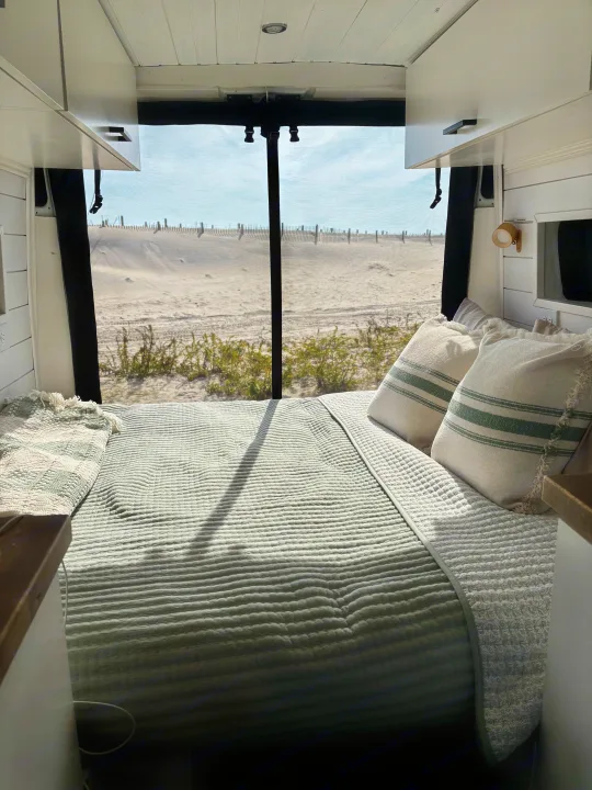 Inside a cozy RV rental: a bed facing a sandy beach view, perfect for van life adventures by the sea.. Ram Ram ProMaster 2500 2019