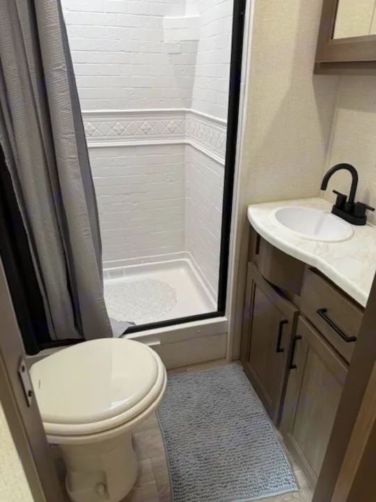 RV rental bathroom features a clean toilet, shower stall with curtain, and sink vanity. Compact & functional space.. Grand Design Transcend 2025