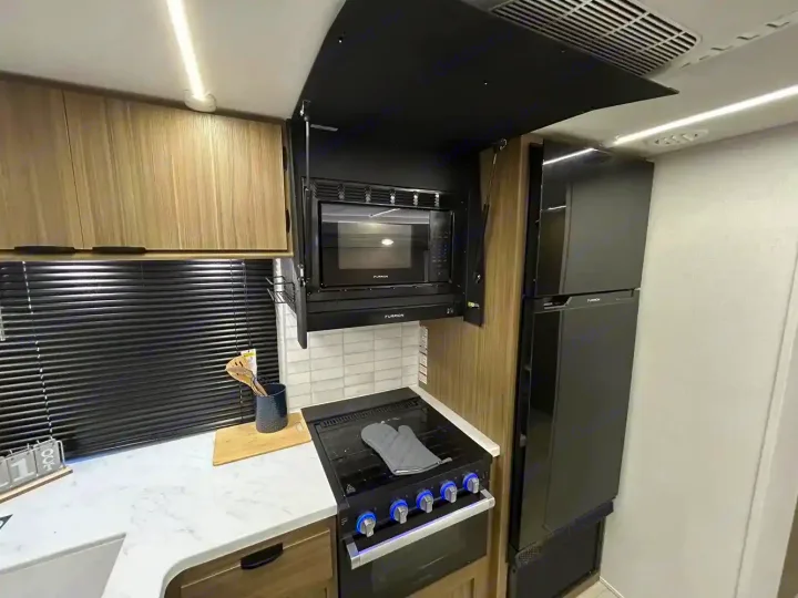 Explore this modern rv rental kitchen with black appliances (microwave, stove, fridge), light wood cabinets, and sleek design.. Forest River Wildwood X-Lite 2025