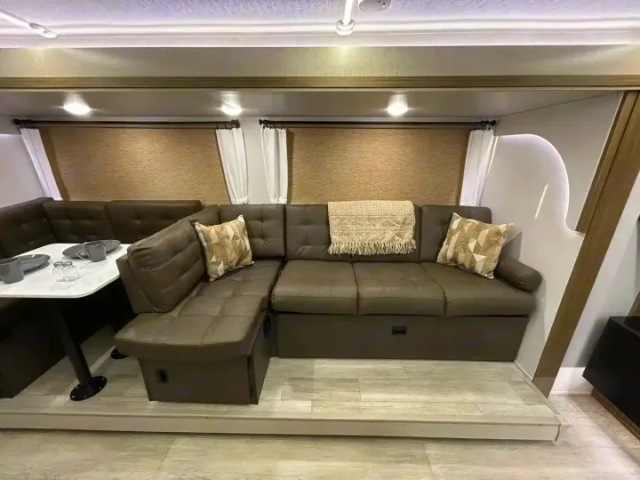 Cozy RV rental interior: large taupe sectional sofa, dinette with table settings, and light wood flooring.. Forest River Wildwood X-Lite 2025