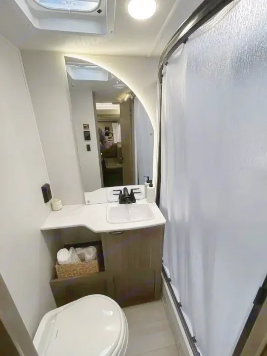 A compact bathroom in an RV rental, showing a toilet, vanity with sink, mirror reflecting another room, and a shower with a curtain.. Forest River Wildwood X-Lite 2025