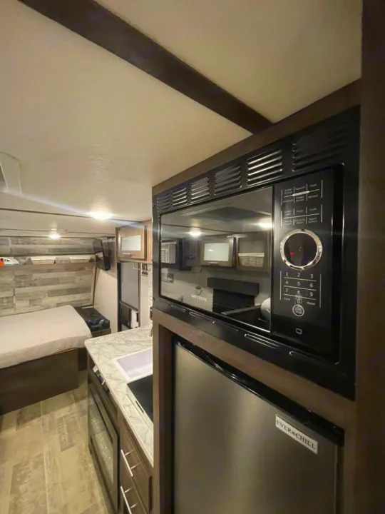 This RV rental features a compact kitchen with a microwave, fridge, and a cozy sleeping area in the background.. LANTERN 17B 2021