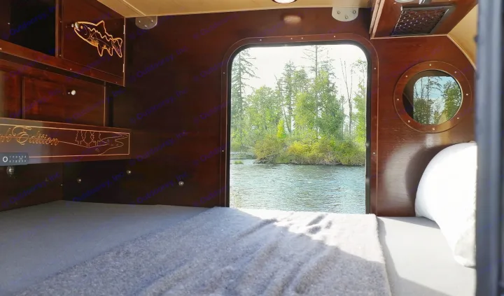 An RV rental's cozy bed looking out onto a peaceful river and forest, offering a tranquil escape into nature.. Teardrops NW Ranger 2025