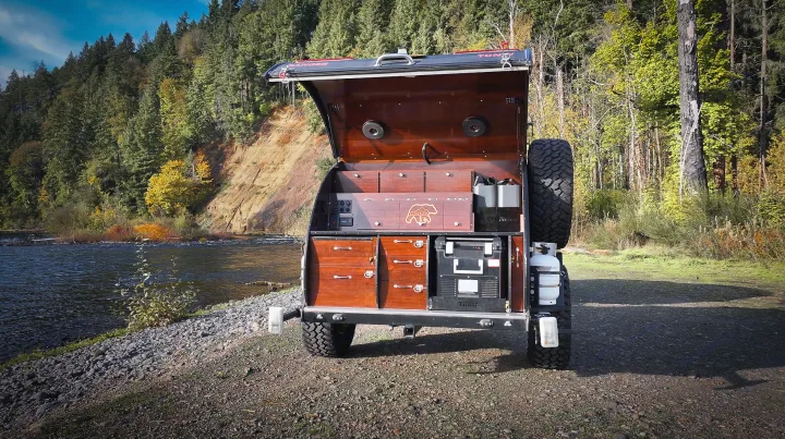 This off-road teardrop camper, with its open galley kitchen revealed, is parked by a scenic river & forest. An ideal RV rental for nature exploration.. Teardrops NW Ranger 2025