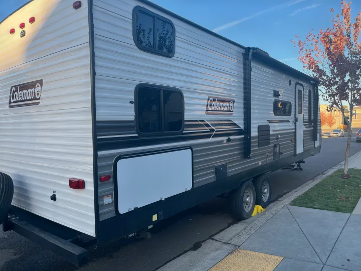 A white and gray Coleman travel trailer RV rental is parked on a residential street under a clear blue sky.. Dutchmen Coleman Lantern 2022
