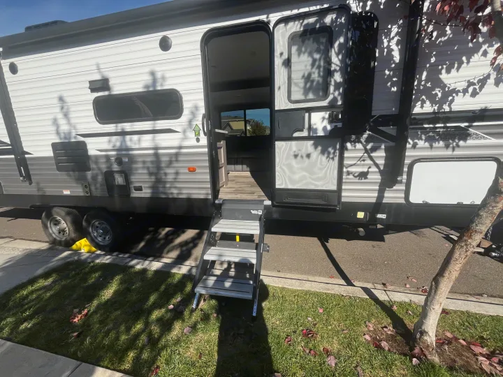 This RV rental is parked with its main entry door open and steps extended, dappled in sunlight and tree shadows.. Dutchmen Coleman Lantern 2022