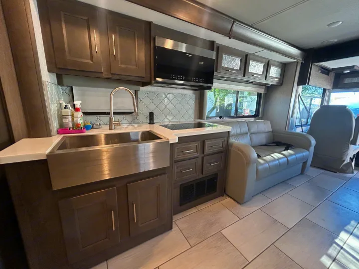 An RV rental interior showing a modern kitchen with a large farmhouse sink and cooktop, adjacent to a comfy sofa and driver's seat.. Forest River Coachmen Sportscoach RD 2021