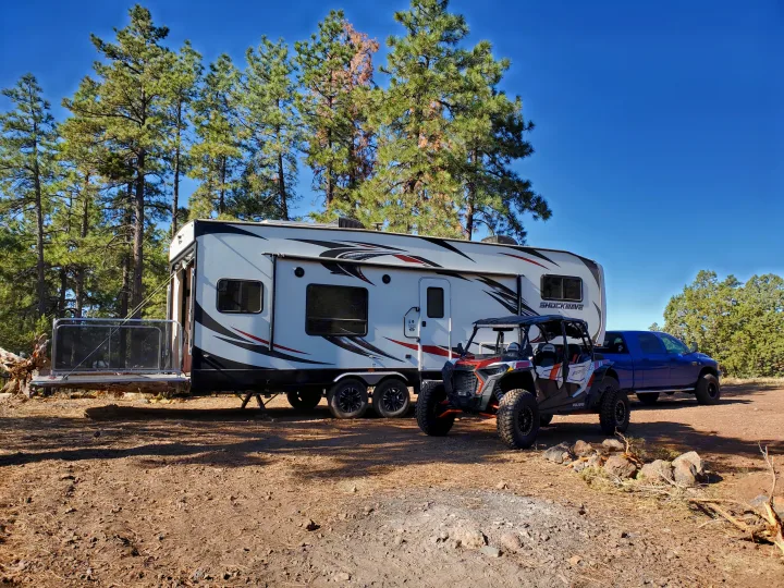 Your next RV rental journey awaits! A spacious travel trailer, UTV, and pickup truck are set up in a pine forest campsite.. Forest River Shockwave 2018
