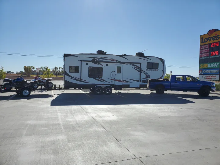 A blue pickup truck tows a large white RV rental and a trailer with ATVs at a Love's gas station on a sunny day.. Forest River Shockwave 2018