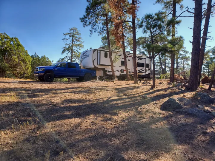 Blue truck with a white fifth-wheel RV (perfect for an RV rental) in a sunny, wooded camp.. Forest River Shockwave 2018