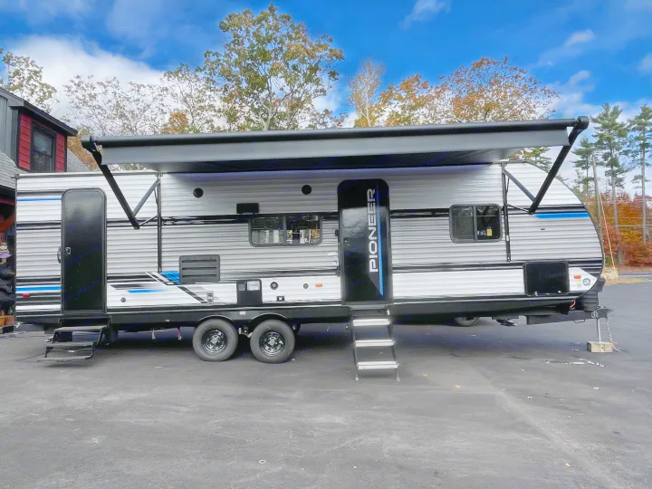 A modern grey and blue travel trailer with an extended awning, parked on asphalt under a blue sky, perfect for an RV rental.. Heartland Pioneer 2024