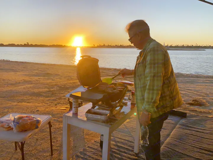 Sunset grilling on a beach by the water, perfect end to an RV rental adventure. Man cooks food on a portable grill.. Thor Outlaw 2017