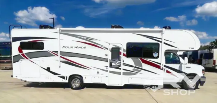 Side view of a white Four Winds Class C RV rental with black, red, and grey graphics, parked outdoors under a blue sky.. Thor Four Winds 2023