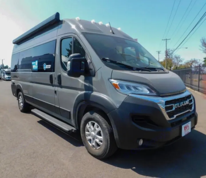 A sleek gray Ram ProMaster camper van for rv rental, parked outdoors on asphalt, featuring a rolled-up awning and side steps.. thor Sequence 2026