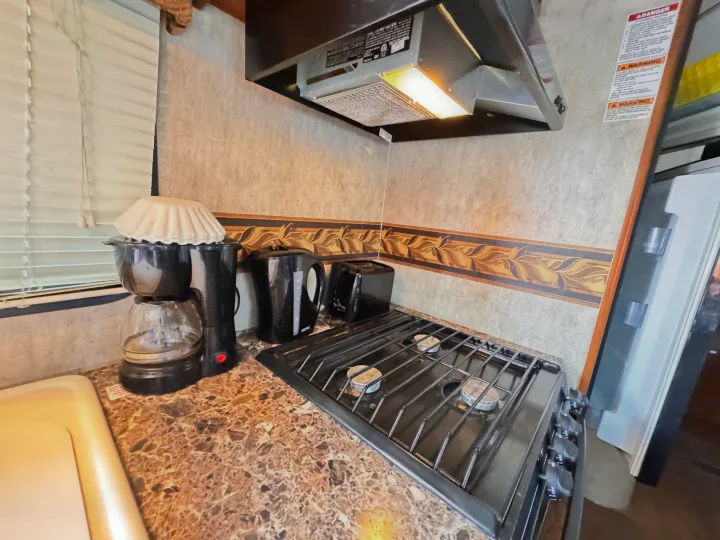 This RV rental kitchen features a coffee maker, kettle, toaster, gas stove, and illuminated range hood.. Keystone RV Bullet Ultra Lite 2014