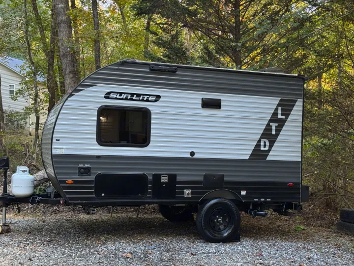 Grey and white Sun Lite LTD travel trailer, an ideal RV rental, parked on a gravel lot surrounded by lush trees.. Sunset Park RV Sun Lite LTD 2024