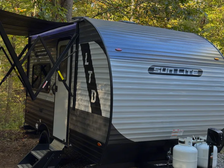 This Sun Lite travel trailer RV rental, with its awning and LED lights extended, is ready for camping in a wooded area.. Sunset Park RV Sun Lite LTD 2024