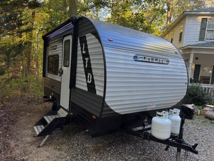 This Sun Lite LTD travel trailer, possibly an RV rental, is parked in a wooded area with its awning extended and propane tanks visible.. Sunset Park RV Sun Lite LTD 2024