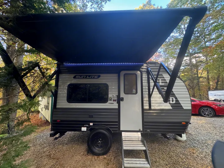 Enjoy this RV rental! A Sun Lite travel trailer with an illuminated awning is set up on gravel in a wooded camp spot.. Sunset Park RV Sun Lite LTD 2024