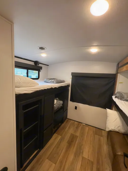 This RV rental bunk room features comfortable beds with white linens, built-in storage, a window with a black shade, and wood-look flooring.. Keystone RV Outback 2022