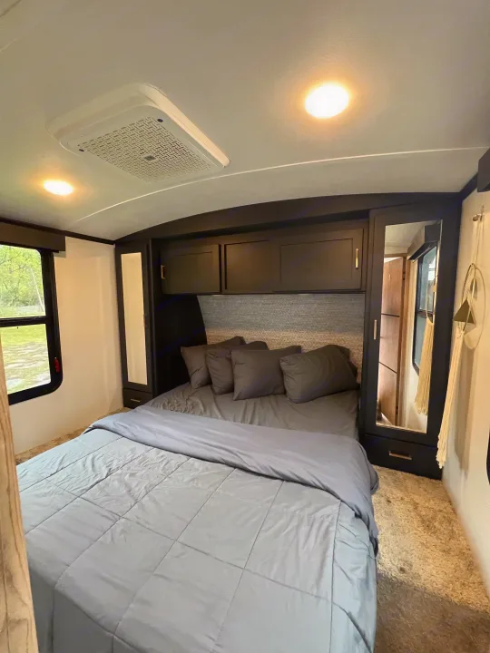 Cozy RV rental bedroom with gray bedding, dark overhead storage, mirrored wardrobe, window, and ceiling AC unit.. Keystone RV Outback 2022