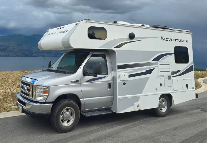 A silver Class C RV rental is parked overlooking a vast lake and mountains under a dramatic, stormy sky.. ALP Adventurer 19RD 2021