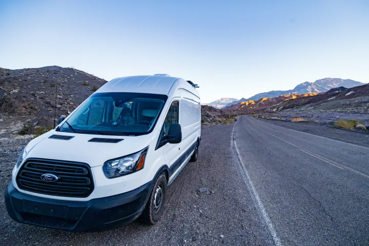 White camper van RV rental parked on a desert road at sunrise/sunset. Mountains glow with golden light in the distance.. Ford Transit 2019