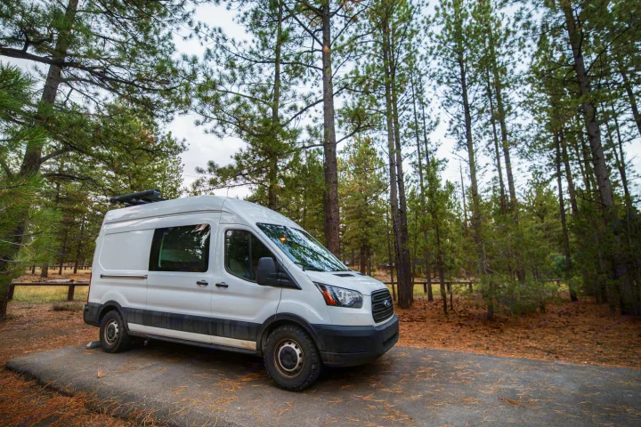 White RV rental van with off-road tires parked among tall pine trees in a peaceful forest setting.. Ford Transit 2019