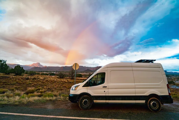 RV rental van parked in a stunning desert landscape with a vibrant rainbow arching across a dramatic sky over mountains.. Ford Transit 2019