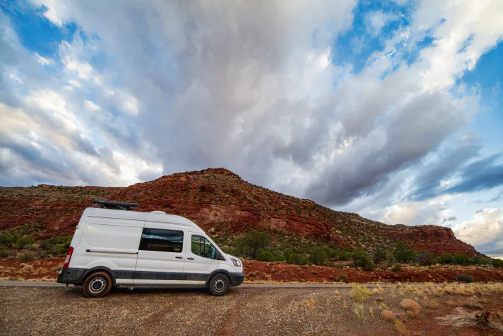 White rv rental van on a desert road with striking red rock mountains and dramatic clouds. Ready for adventure.. Ford Transit 2019