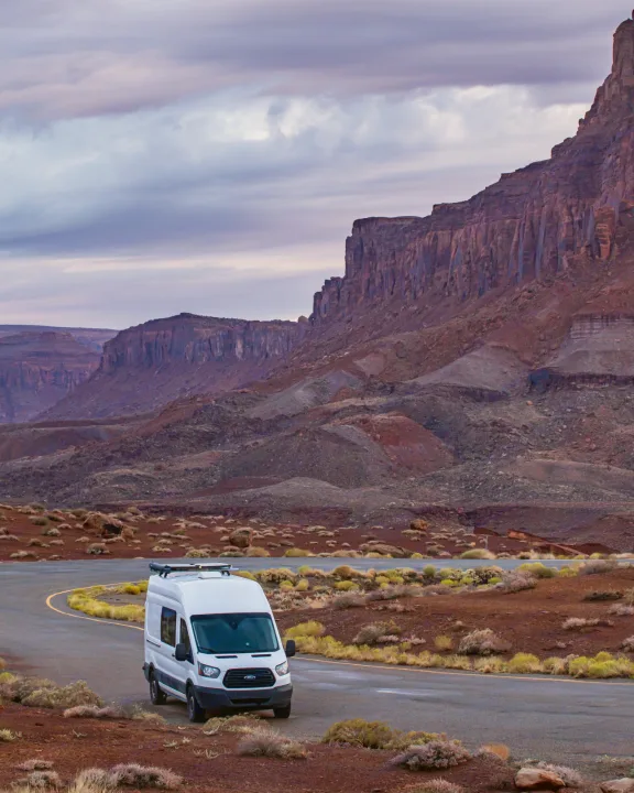 White rv rental campervan parked on a winding desert road, surrounded by majestic red cliffs and a dramatic sky.. Ford Transit 2019