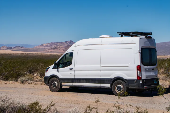 White high-roof RV rental van, off-road ready, parked on a desert dirt path with mountains under a blue sky.. Ford Transit 2019