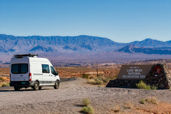 An RV rental van pauses at the entrance to Lake Mead National Recreation Area, with vast desert mountains behind.. Ford Transit 2019