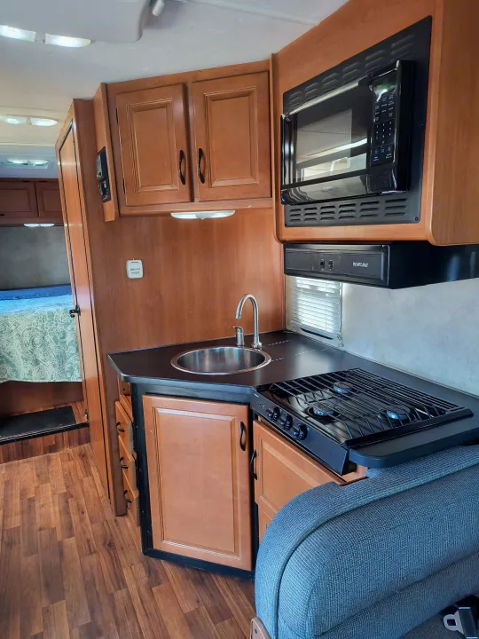 Compact RV rental kitchen featuring a microwave, stove, sink, wood cabinets, and glimpse of bed/seating.. Thor Motor Coach Four Winds Majestic 2014