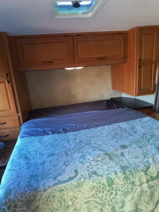 Cozy rv rental bedroom featuring a bed with patterned blue blanket, overhead wood cabinets, ceiling vent, and side storage.. Thor Motor Coach Four Winds Majestic 2014