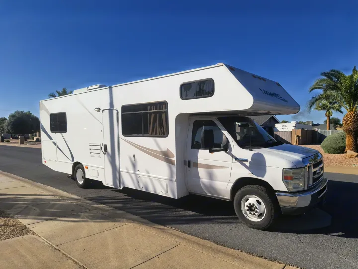 A sleek white Class C RV rental with tan accents is parked on a sunny street. Blue sky above, palm trees in the background.. Thor Motor Coach Four Winds Majestic 2014