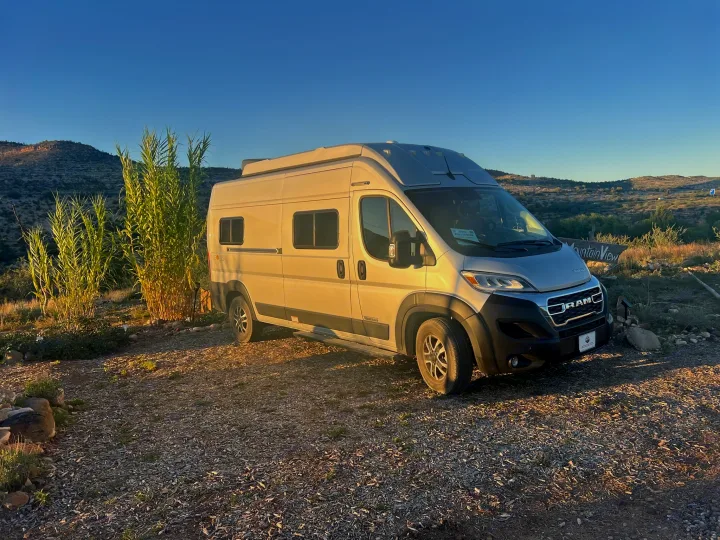 This silver camper van rv rental is parked in golden light, with tall plants & distant mountains under a blue sky.. Winnebago Soilis 2025