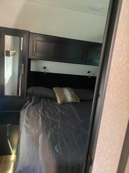 This cozy RV rental bedroom features a comfy bed with dark gray bedding, overhead cabinets, and a mirrored closet.. Forest River Cherokee Grey Wolf 2022