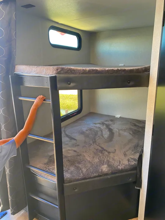 Cozy RV rental bunk beds with windows, perfect for family travel. A child's arm is seen on the ladder to the top bunk.. Forest River Cherokee Grey Wolf 2022