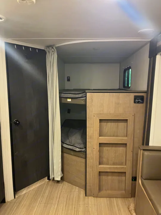 RV rental interior featuring cozy bunk beds with nets, a dark door, privacy curtain, and light wood finishes.. Keystone RV Hideout 2021